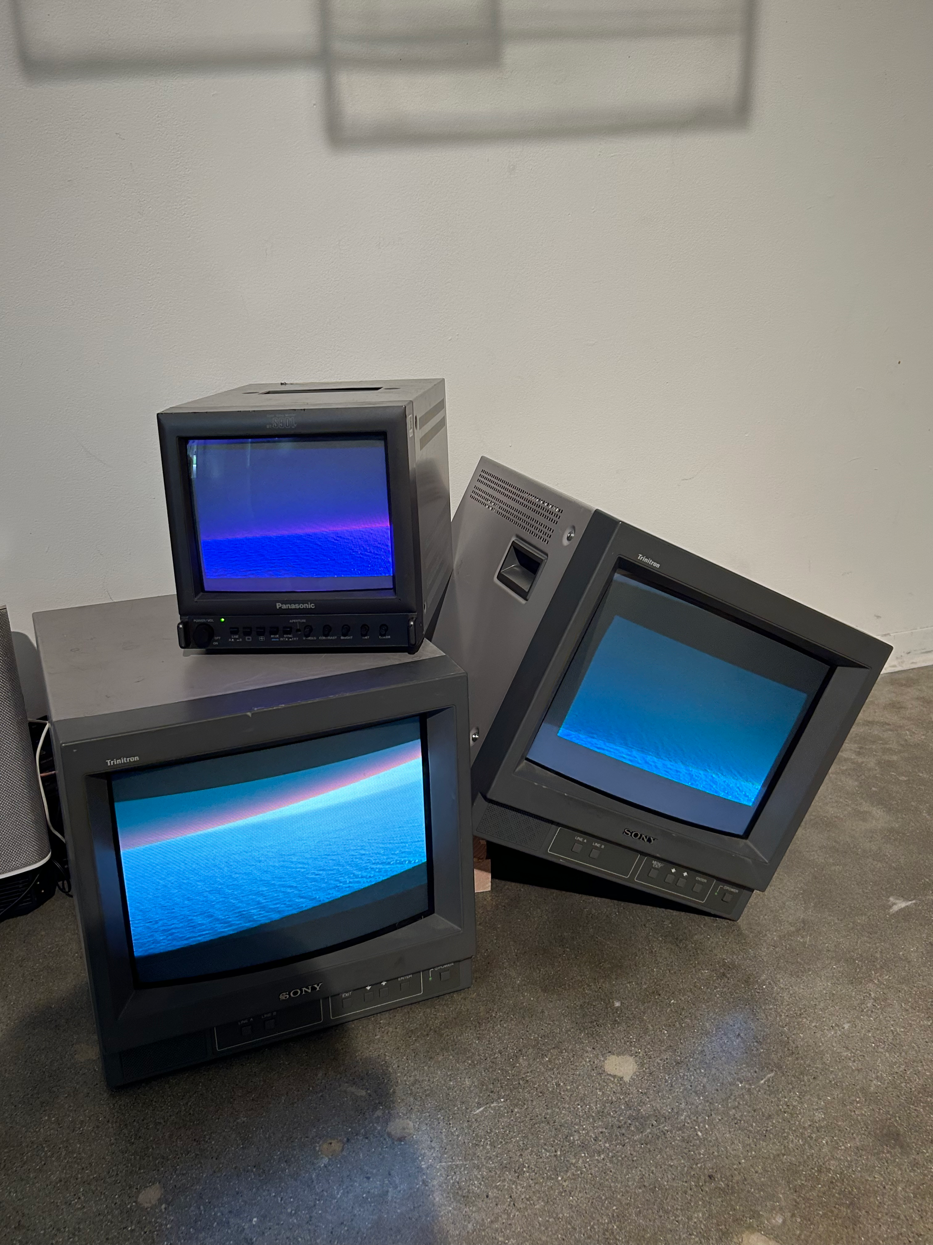 On the floor of the gallery three CRT TV's sit, one leaning against the other, and another placed on top.