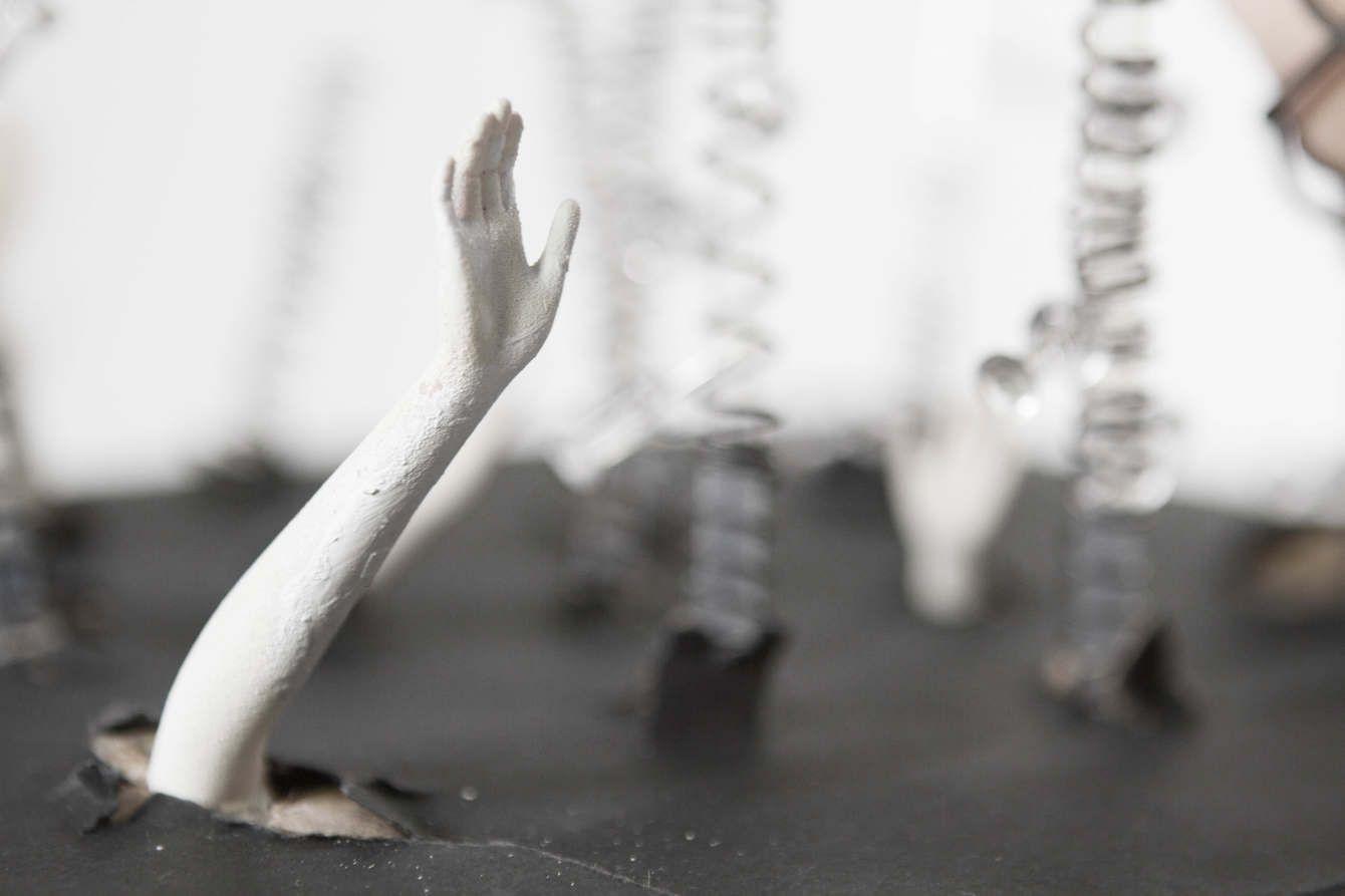 There is a doll hand painted white coming out of a black material. In the background there are blurry clear rods with wire wrapped around them.