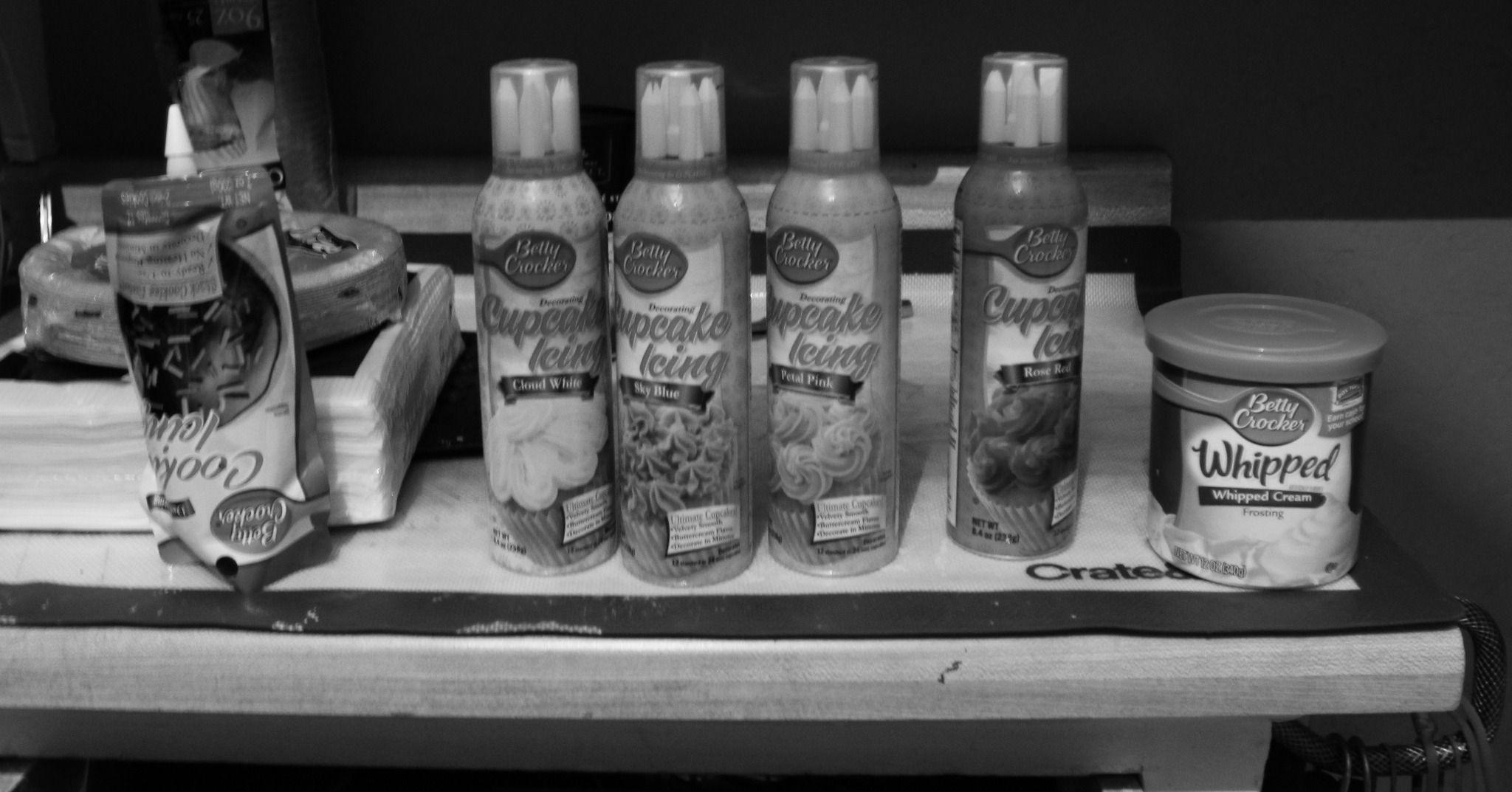 There is a photo of a table with various frostings and icings on it, all from the Betty Crocker brand. There are four cans of Cupcake Icing (Cloud White, Sky Blue, Petal Pink, Rose Red), Whipped Cream frosting, and an upside down frosting bag.